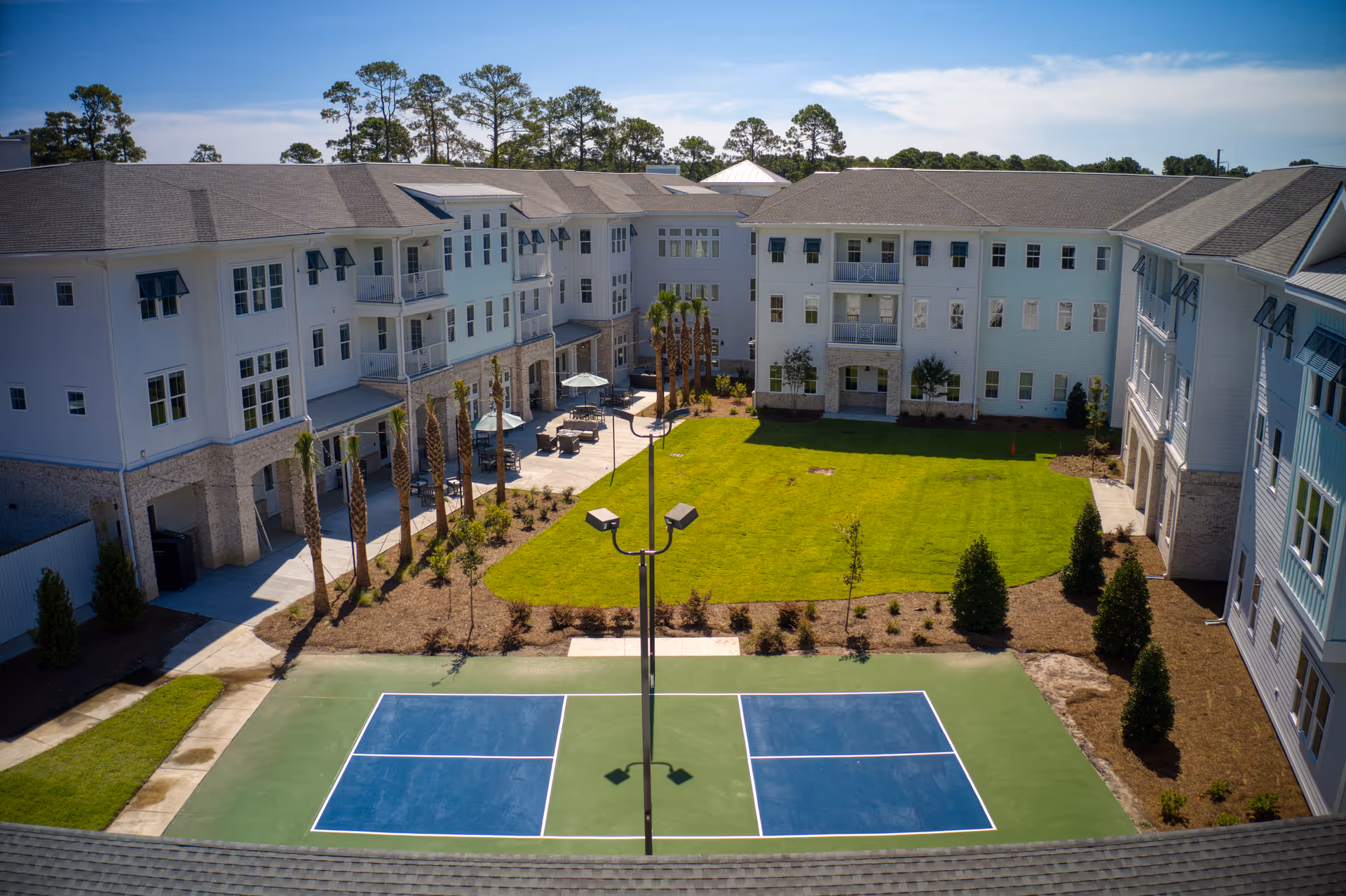 Courtyard view of a senior living complex with a central green lawn, patio seating, palm trees and a blue sports court surrounded by three-story white buildings.