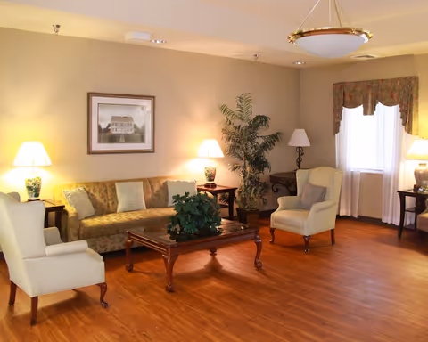 A cozy living room with wooden flooring, featuring a patterned sofa with cushions, two cream-colored armchairs, a wooden coffee table with a green plant centerpiece, and several lamps providing warm lighting. There is a framed picture on the wall and a large potted plant in the corner near a window with floral curtains.