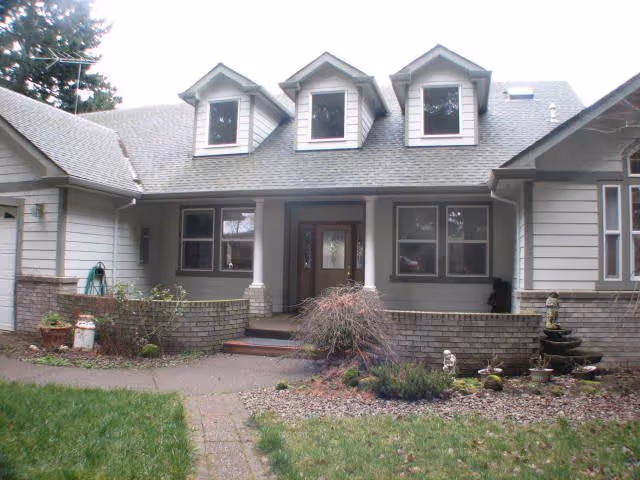 Front exterior view of a single-story house with three dormer windows on the roof, a covered porch with two white columns, a wooden front door with glass panels, and a small garden area with plants and shrubs in front.