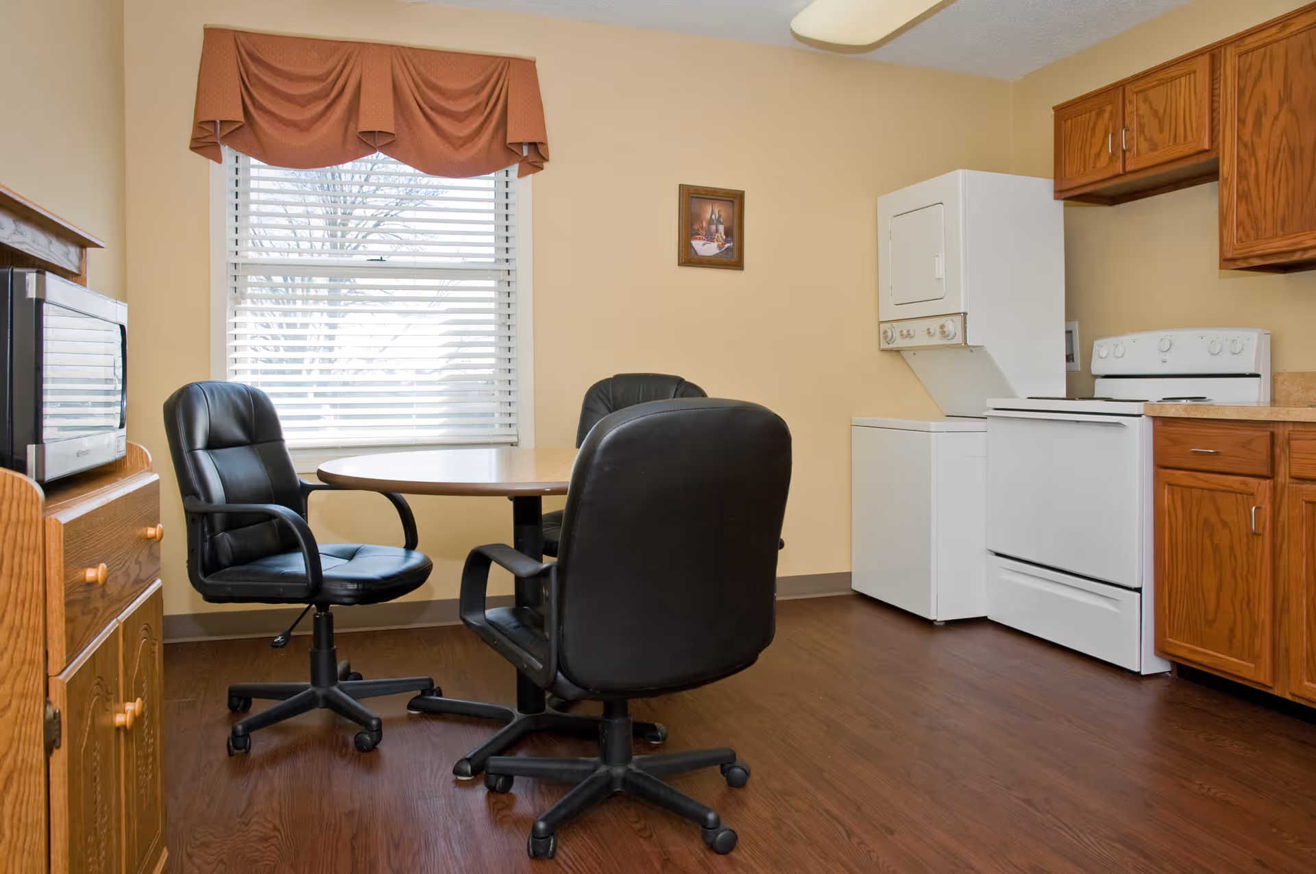 A small kitchen and dining area with a round wooden table surrounded by three black office chairs on wheels. The kitchen features wooden cabinets, a white stove, a stacked washer and dryer unit, and a microwave on a wooden cabinet. A window with white blinds and a brown valance is visible behind the table.