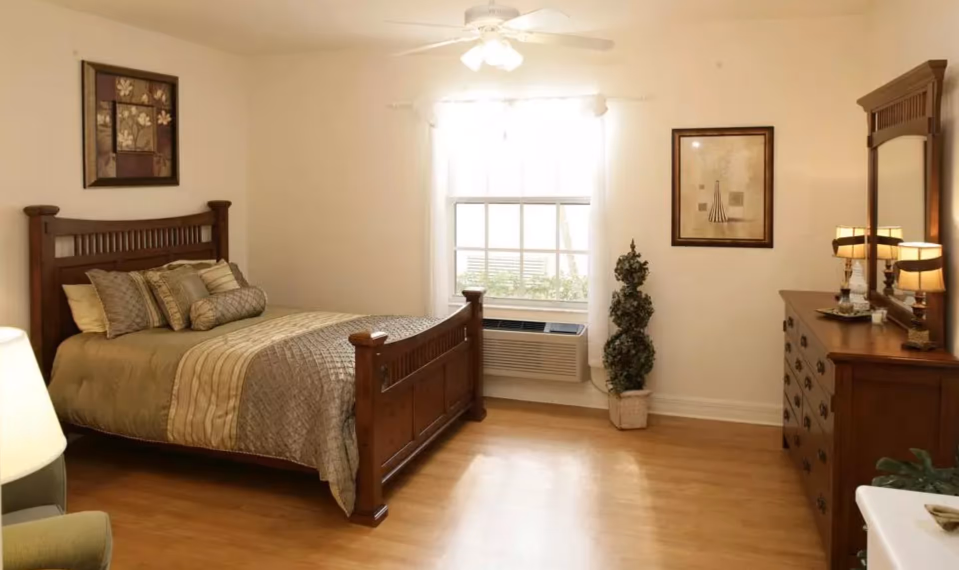 Sunlit bedroom with a wooden bed and matching dresser, a window with an air conditioner, and hardwood floors.