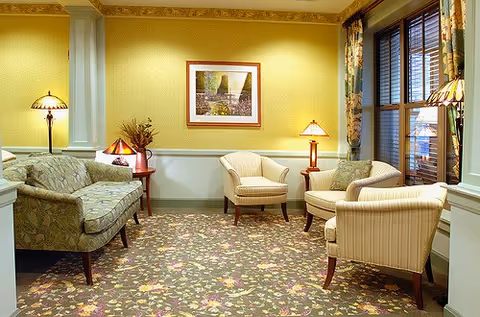 A cozy sitting area with floral patterned carpet, a green floral sofa, three beige armchairs, two side tables with lamps, a framed picture on a yellow wall, and a window with floral curtains.