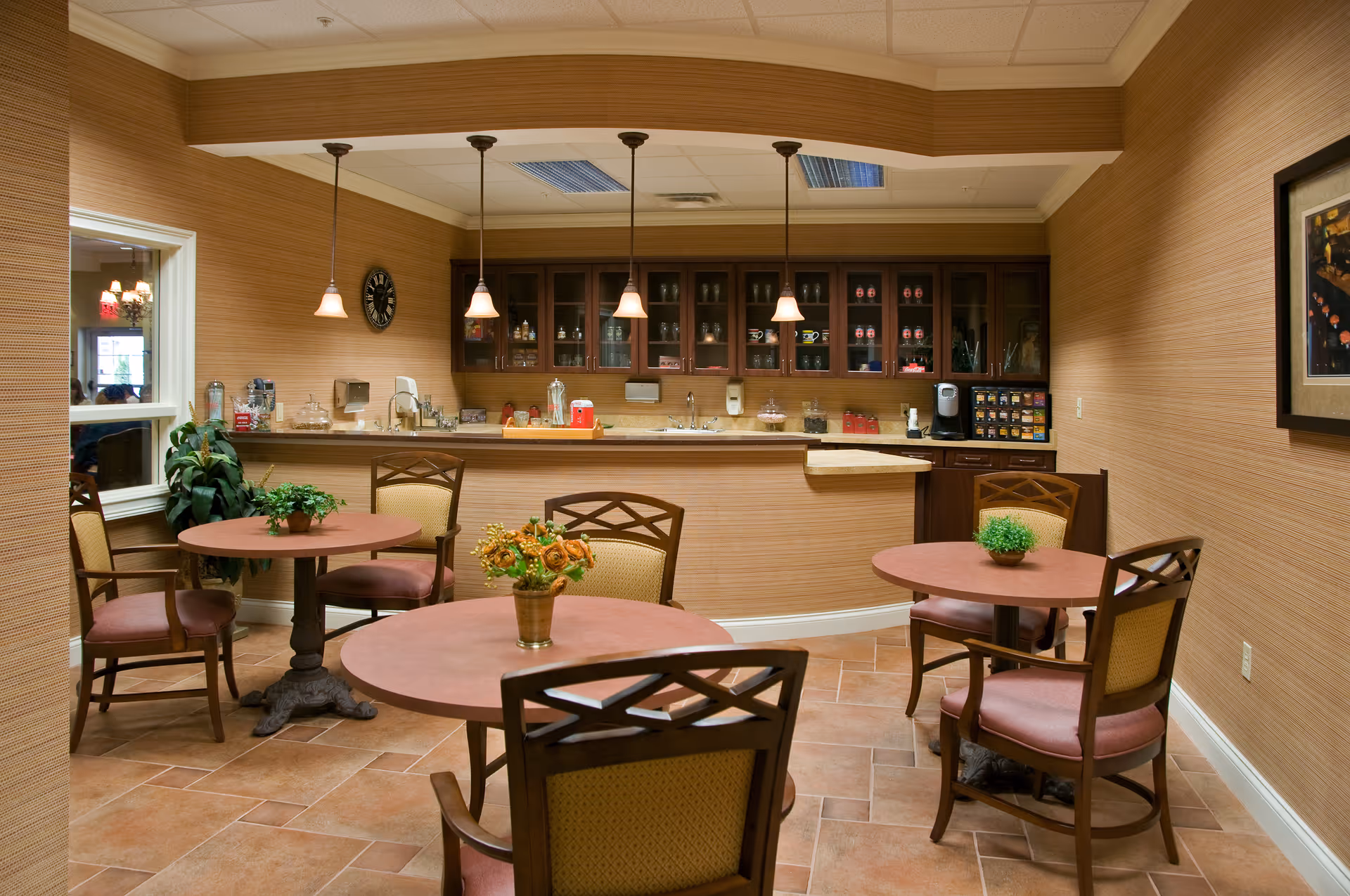 A cozy dining area with three round tables, each surrounded by four cushioned wooden chairs. The tables have small decorative plants or flower arrangements. In the background, there is a counter with a sink and cabinets filled with glassware and other items. The walls are covered with a textured beige wallpaper, and the floor is tiled. Pendant lights hang from the ceiling above the counter, and a framed picture is visible on the right wall.