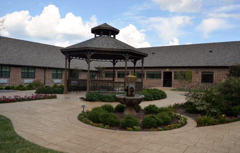 Outdoor courtyard area of a senior living facility with a central gazebo, a water fountain surrounded by bushes and flowers, paved walkways, and a brick building with multiple windows in the background under a partly cloudy sky.
