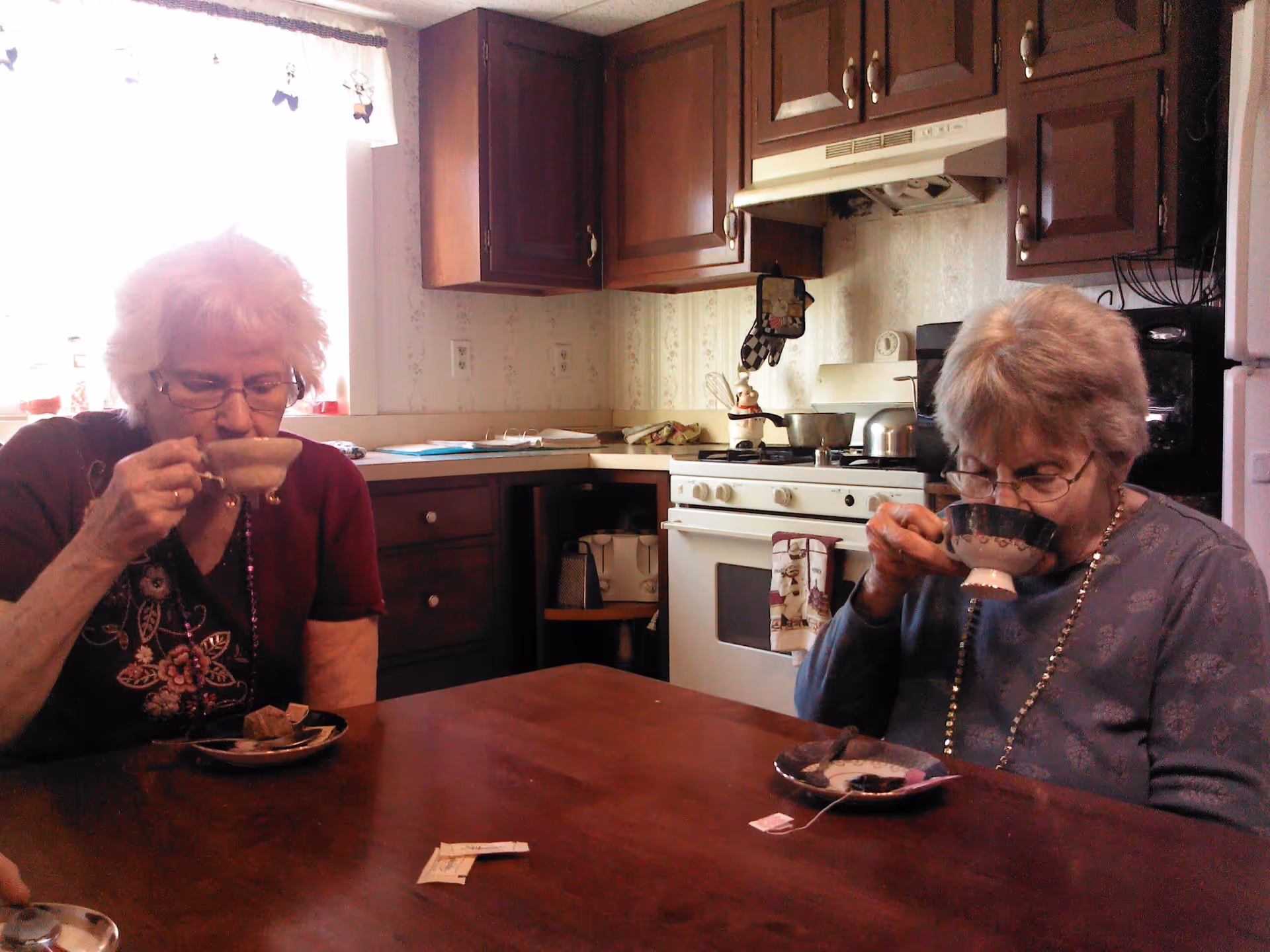 Two elderly women sitting at a wooden table in a kitchen, each drinking from a teacup. The kitchen has dark wooden cabinets, a white stove with a tea kettle on it, and a window with a white curtain letting in natural light.