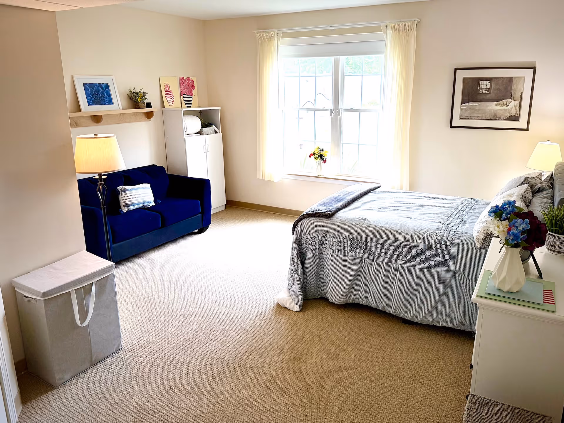 Bright furnished bedroom with a bed and nightstand, a blue loveseat, window with curtains, and light carpeting.