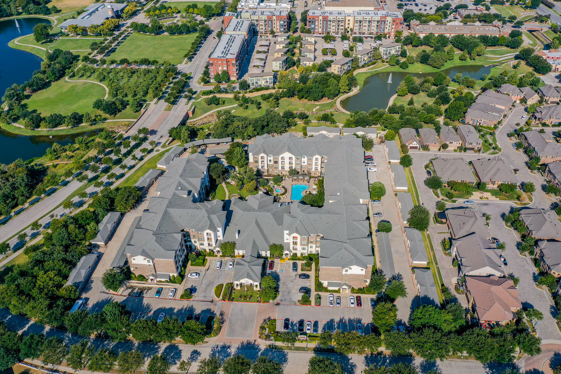 Aerial view of Conservatory At Keller Town Center senior living facility showing multiple buildings with gray roofs surrounding a central courtyard with a swimming pool and hot tub. The facility is bordered by parking lots, roads, green lawns, trees, and nearby residential neighborhoods. A small pond with a fountain is visible in the background.
