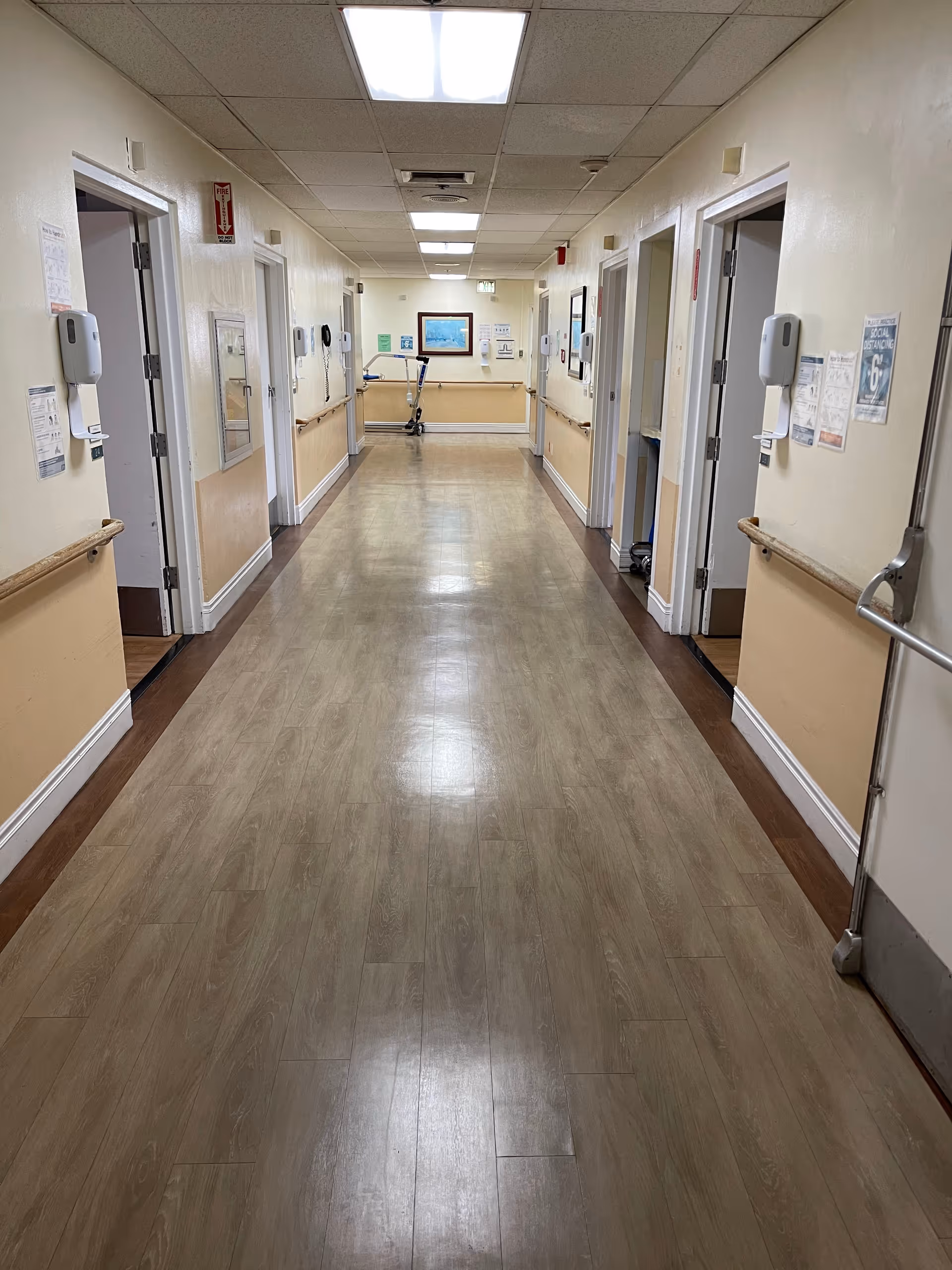 Long, well-lit hallway in a care facility with handrails along the walls and a walker at the far end.