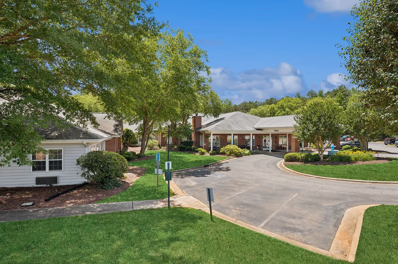 Exterior view of Summit Place of Kings Mountain showing a single-story brick building with a covered entrance driveway, surrounded by green lawns, trees, and shrubs under a blue sky with some clouds.