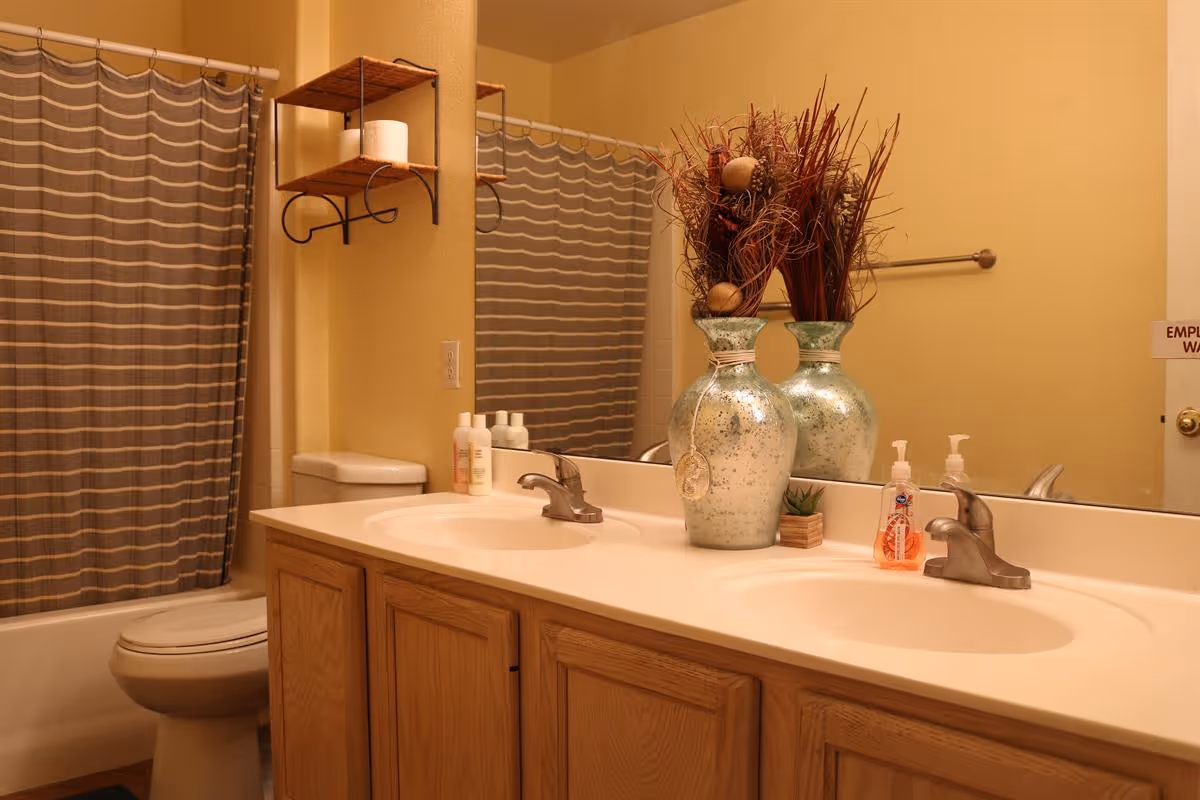 Double-sink bathroom vanity with decorative vases and soap dispensers, mirror, striped shower curtain and toilet in the background.