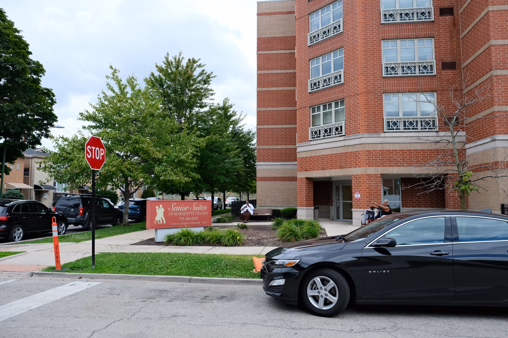 Front entrance of a red-brick Senior Suites of Marquette Village building with its sign, parked cars, and a stop sign on the street.
