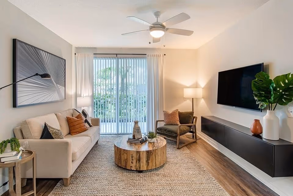 A modern living room with a beige sofa adorned with cushions, a round wooden coffee table, a single armchair with cushions, a wall-mounted flat-screen TV, a floating black media console, a floor lamp, a ceiling fan with light, and large sliding glass doors with vertical blinds leading outside.