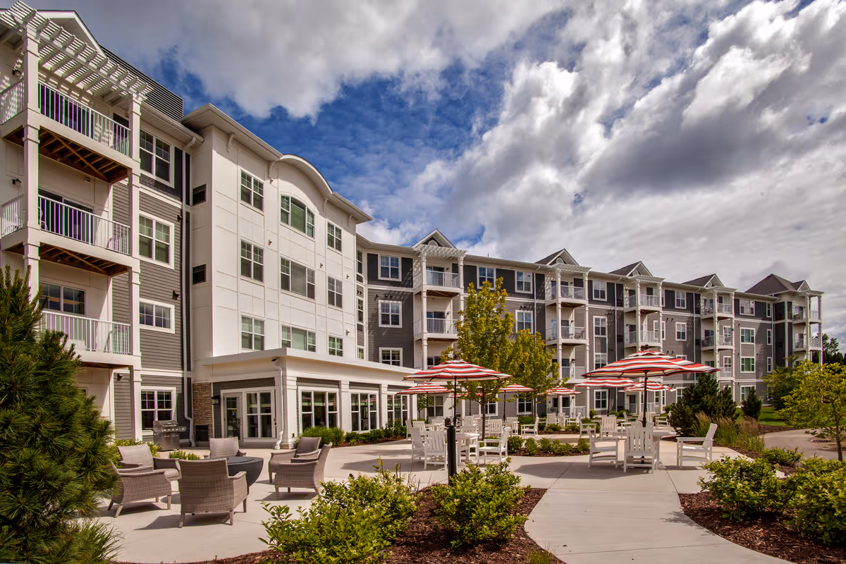 Outdoor patio area of a senior living facility with several seating arrangements including chairs and tables with red and white striped umbrellas. The multi-story building in the background has balconies and large windows under a partly cloudy sky.