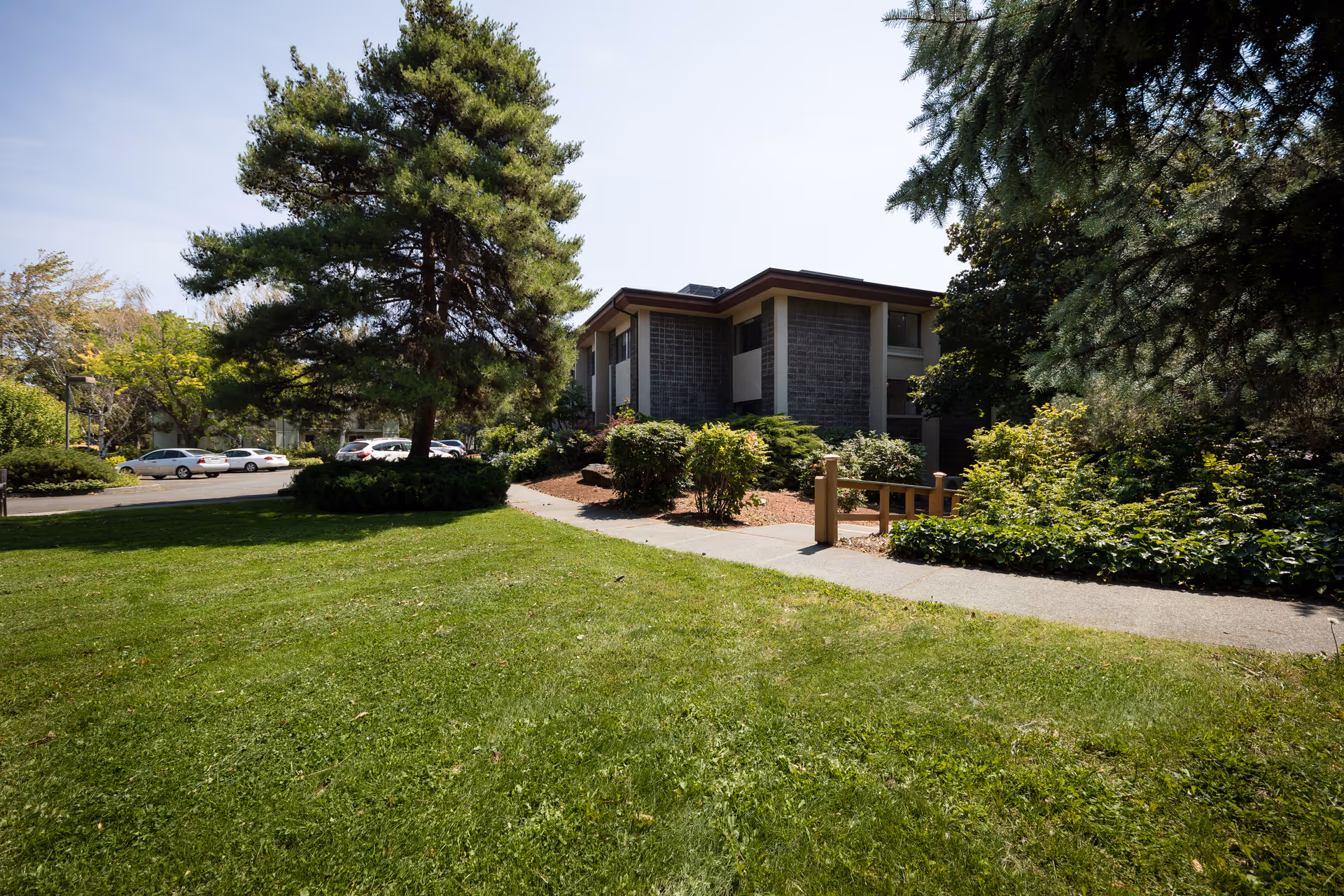 A two-story building surrounded by lush greenery, including large trees, bushes, and a well-maintained lawn. A paved walkway leads to the entrance of the building, and several parked cars are visible in the background under a clear sky.