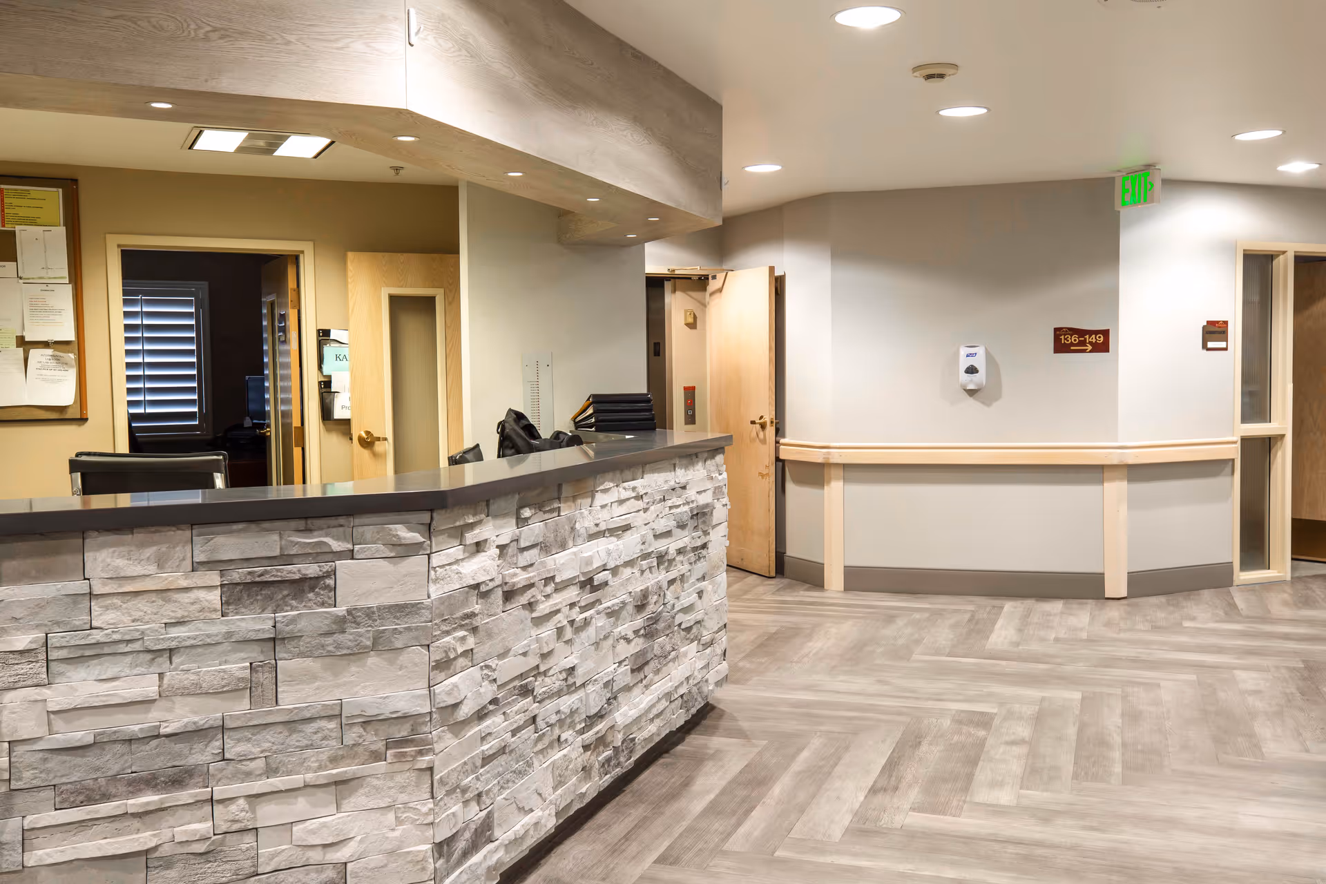 Interior view of a reception or nurse station area in a senior living facility with a stone-faced counter, light wood accents, and a hallway leading to rooms. The floor has a light wood pattern, and there is a hand sanitizer dispenser mounted on the wall.