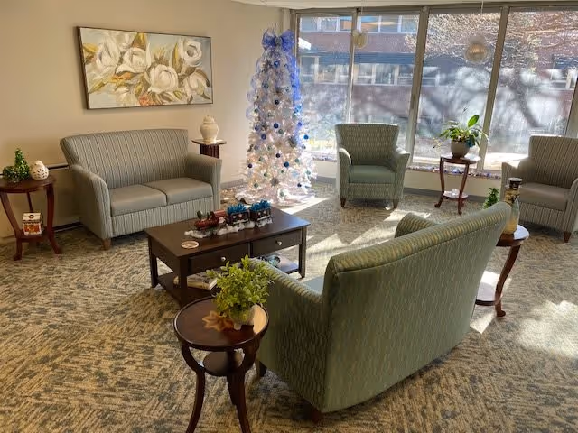 A cozy living room area with light green upholstered sofas and armchairs arranged around a wooden coffee table. There is a white Christmas tree decorated with blue and silver ornaments near a large window letting in natural light. Small side tables with plants and decorative items are placed around the seating area, and a floral painting hangs on the wall.