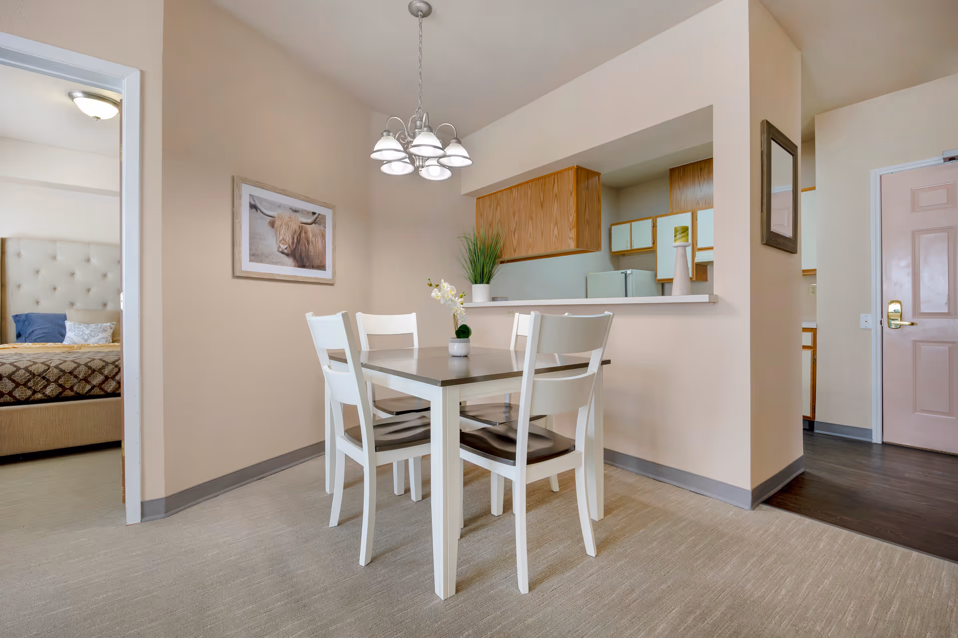 A dining area with a white table and four chairs, a small plant centerpiece on the table, a hanging light fixture above, and a view into a bedroom with a bed and a framed picture of a highland cow on the wall. The dining area is adjacent to a kitchen with wooden cabinets and a refrigerator, and there is a door visible in the background.