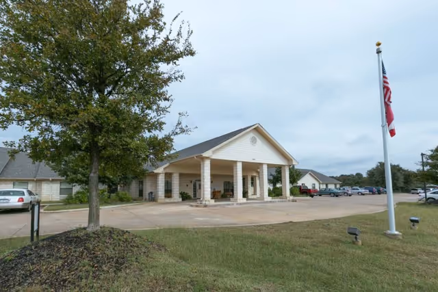 Front entrance of a single-story health center building with a covered porte-cochere, flagpole, tree, and parking lot.