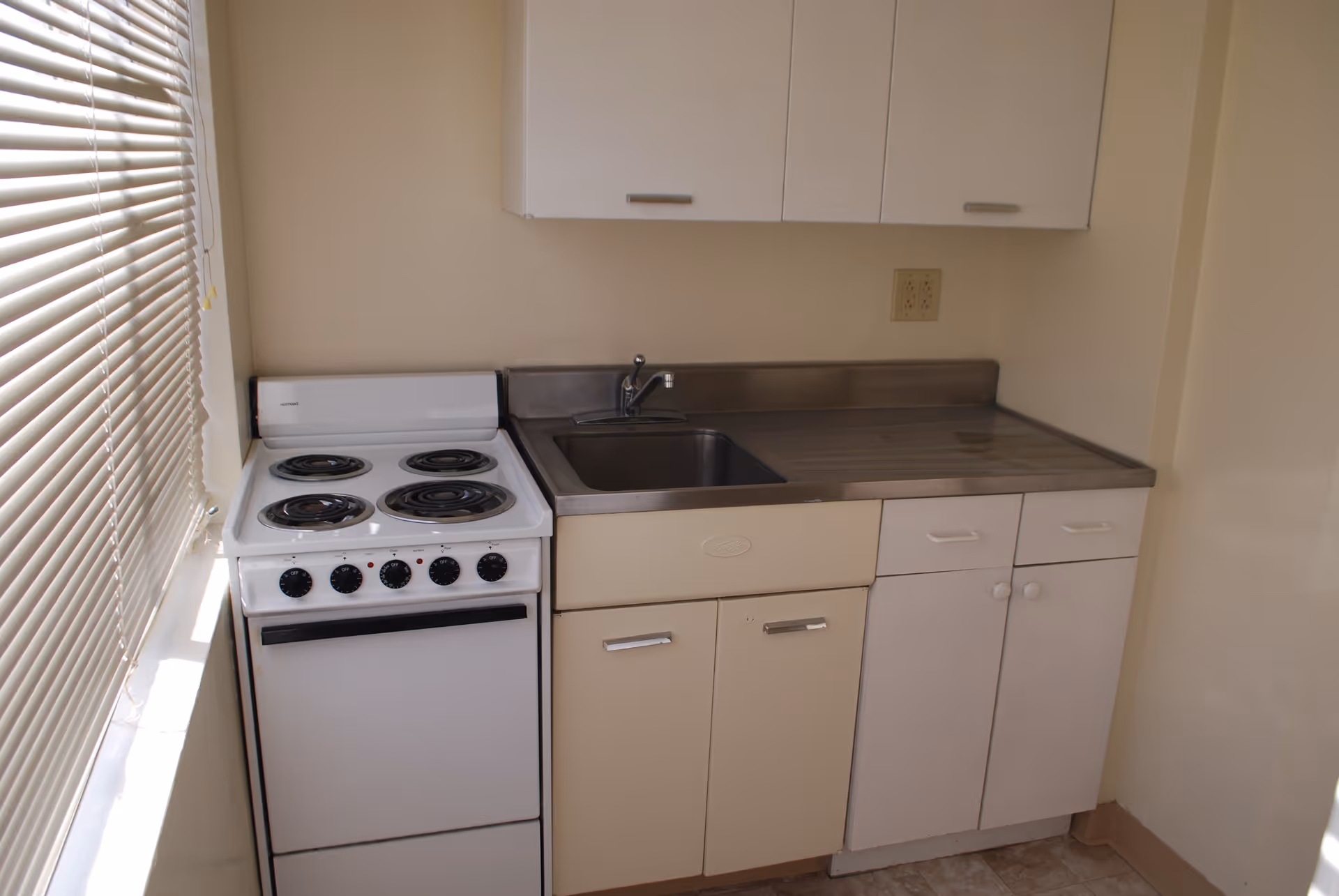 Small kitchen area with a white electric stove with four coil burners on the left, a stainless steel sink in the middle, and white cabinets above and below the countertop. A window with closed blinds is on the left side, allowing natural light into the space.