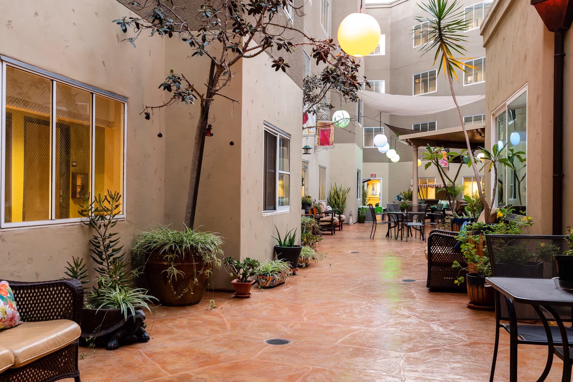 A bright indoor courtyard area in a senior living facility with potted plants, trees, and outdoor furniture including chairs and tables. The space is surrounded by beige walls with windows and has hanging spherical lights overhead.