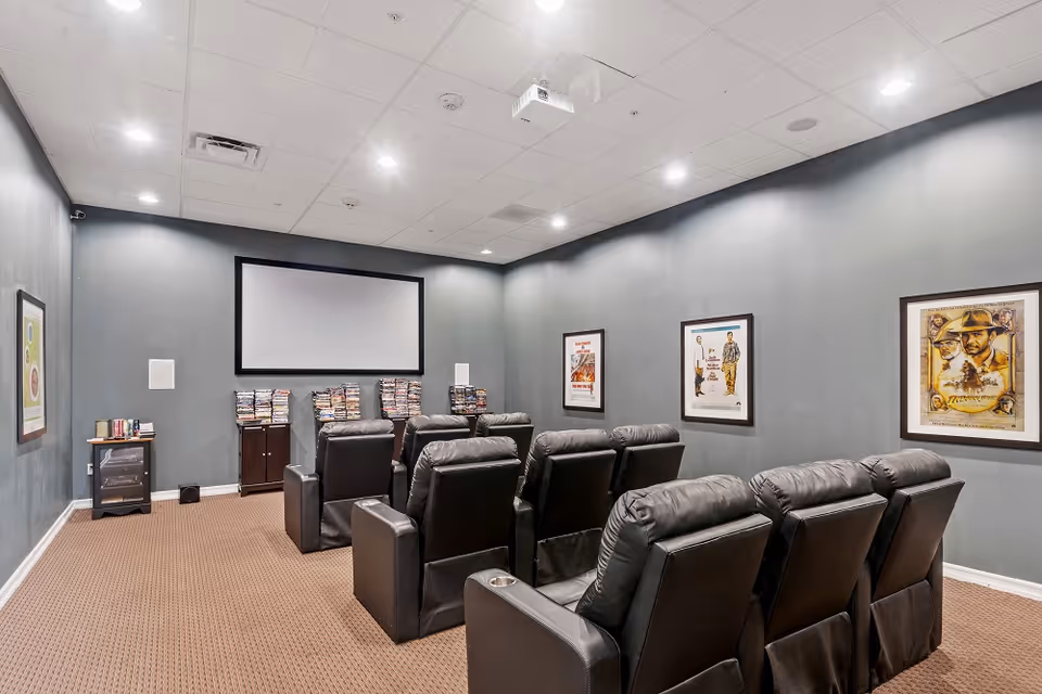 Private screening room with rows of black leather recliners facing a blank projection screen and framed movie posters on gray walls.