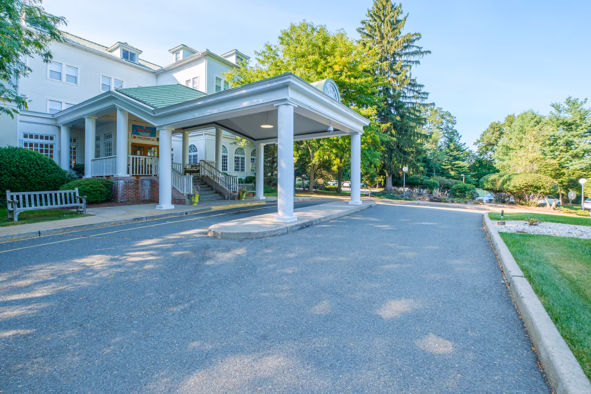 Entrance to a senior living and rehabilitation facility with a covered drop-off area supported by white columns, a green roof, and a multi-story white building in the background surrounded by trees and greenery.