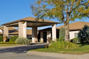 Exterior view of a single-story building with a covered entrance, surrounded by trees and well-maintained landscaping under a clear blue sky.