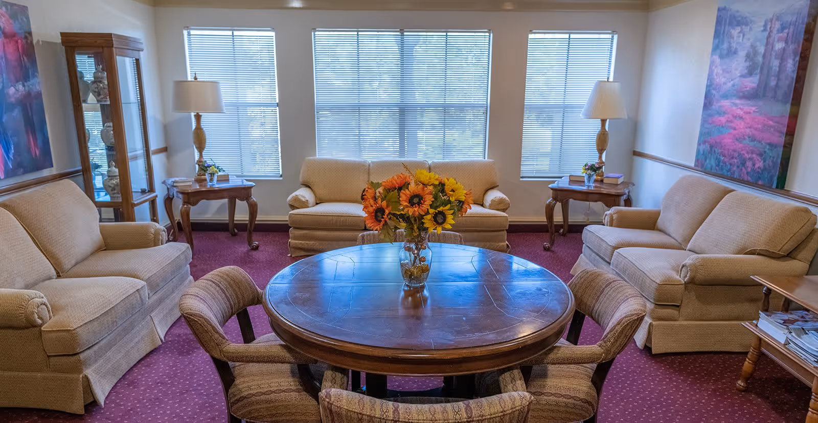 A cozy living room with three beige sofas arranged around a round wooden table with a vase of sunflowers. The room has three large windows with blinds, two wooden side tables with lamps, a glass display cabinet, and colorful paintings on the walls. The carpet is a deep red color.