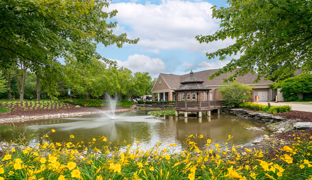 A serene outdoor scene at Autumn House Bloomfield Hills featuring a pond with a water fountain and a wooden gazebo on stilts. The pond is surrounded by lush green trees and vibrant yellow flowers in the foreground. A building with a sloped roof is visible in the background under a partly cloudy sky.