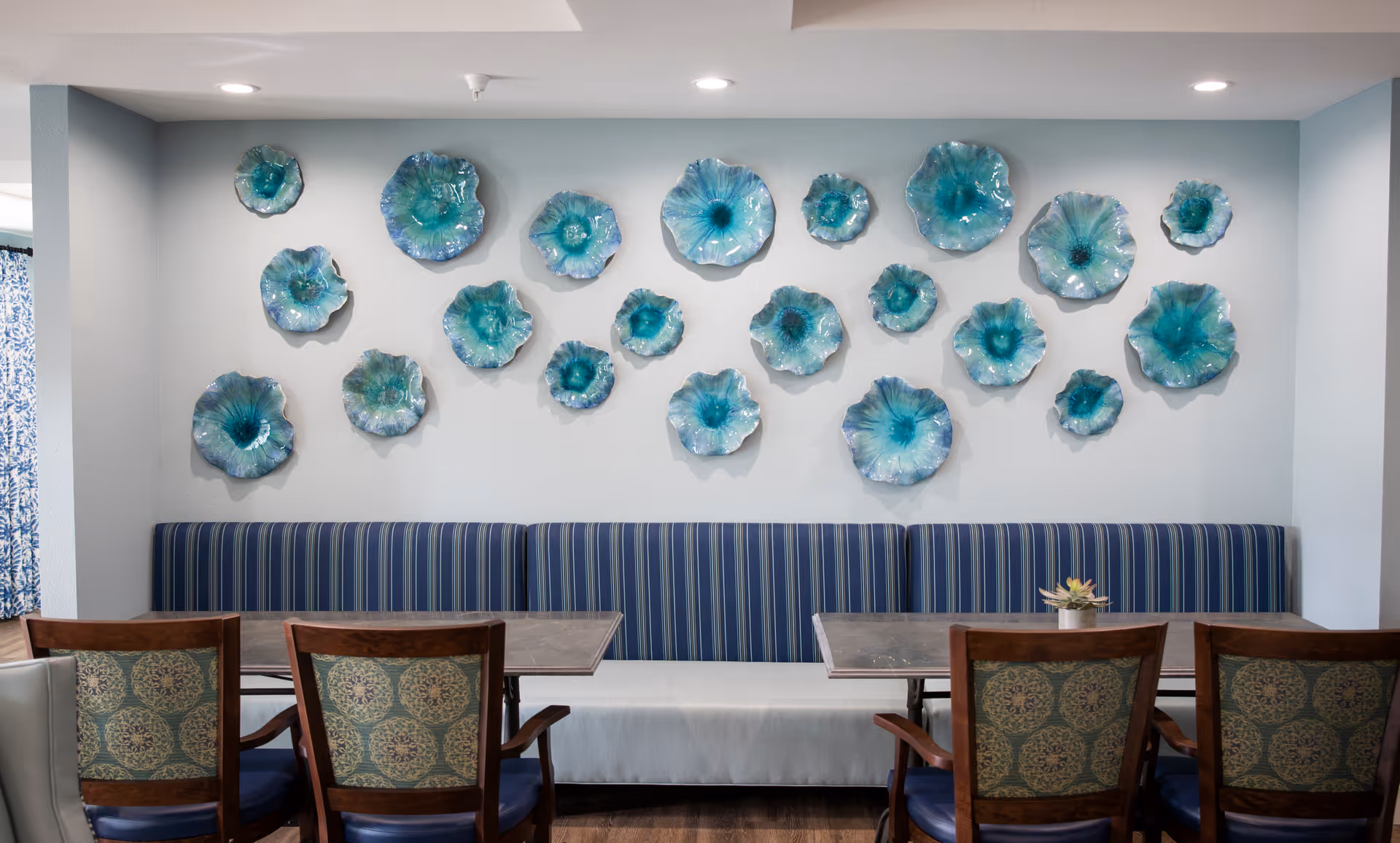 Dining area with striped banquette seating, wooden chairs and tables, and a wall display of blue-green glass flower sculptures.