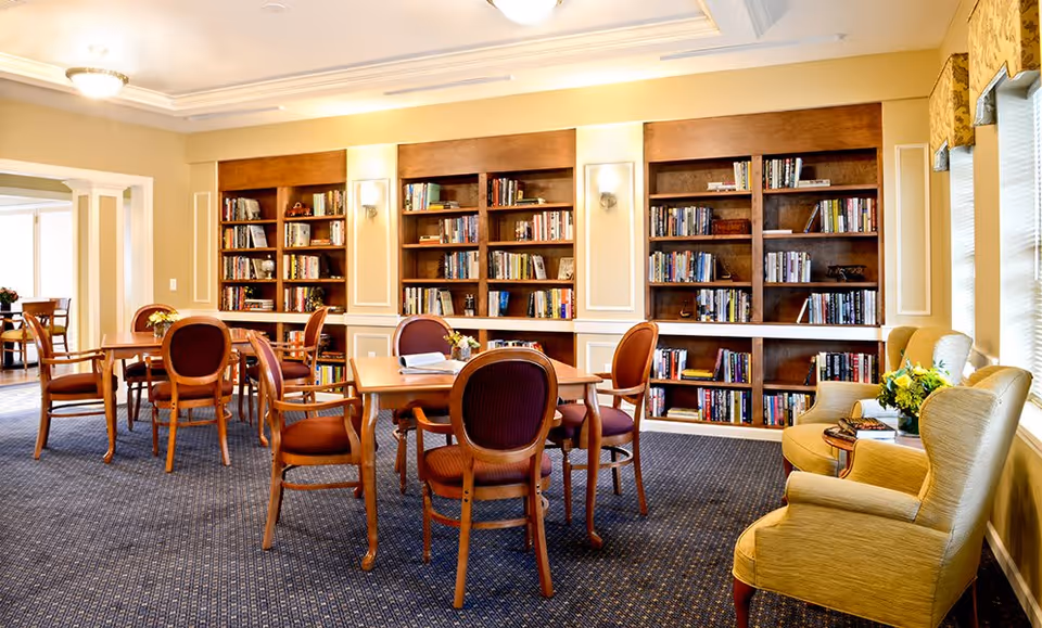 Communal library-style room with wooden bookshelves, tables and chairs, and armchairs by the windows.