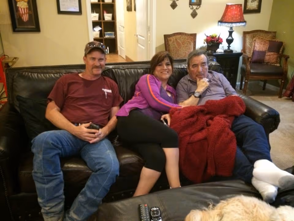 Three people sitting closely together on a dark leather couch in a cozy living room. The person on the left is a man wearing a maroon shirt and blue jeans, the person in the middle is a woman in a purple jacket and black pants, and the person on the right is an older man covered with a red blanket, wearing a gray shirt and white socks. Behind them are chairs, a side table with a lamp, and some decorative items on the walls.