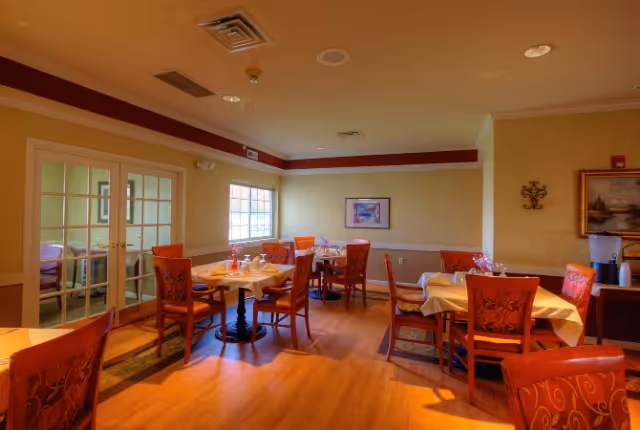 A dining room in a senior living facility with several tables covered with white tablecloths and set with glasses and napkins. The room has wooden flooring, red cushioned chairs with patterns, a window letting in natural light, framed artwork on the walls, and double glass doors leading to another room.