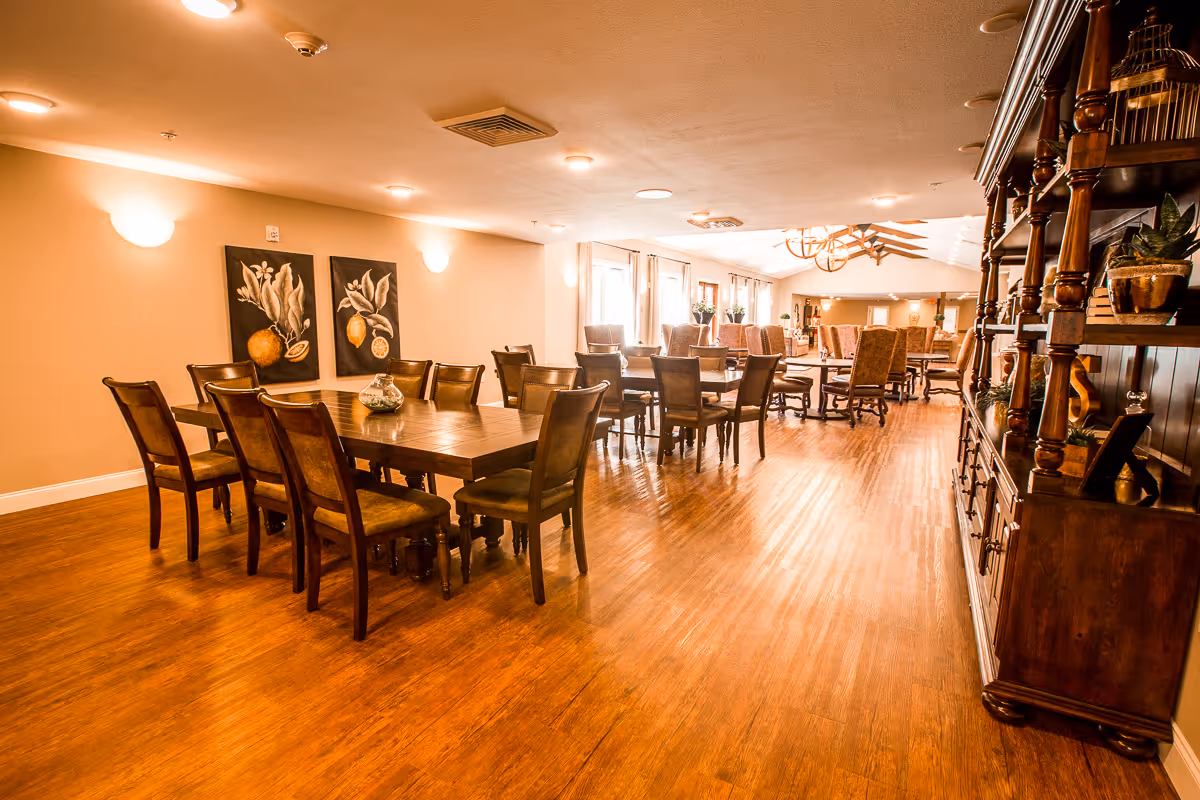 A spacious dining room with multiple wooden tables and chairs arranged neatly. The room has warm lighting, wooden flooring, and decorative paintings on the wall. There is a large wooden cabinet on the right side with plants and decorative items. Windows along the far wall allow natural light to enter the room.
