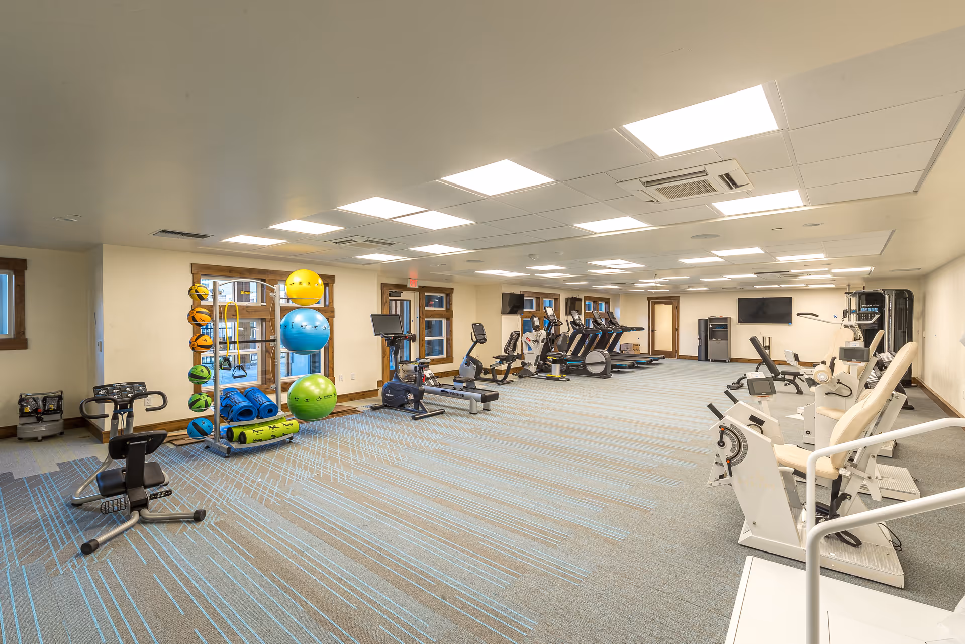 A spacious fitness room with various exercise equipment including stationary bikes, treadmills, and weight machines. There are colorful exercise balls and mats stored on racks near windows with wooden frames. The room has bright overhead lighting and a patterned carpet floor.