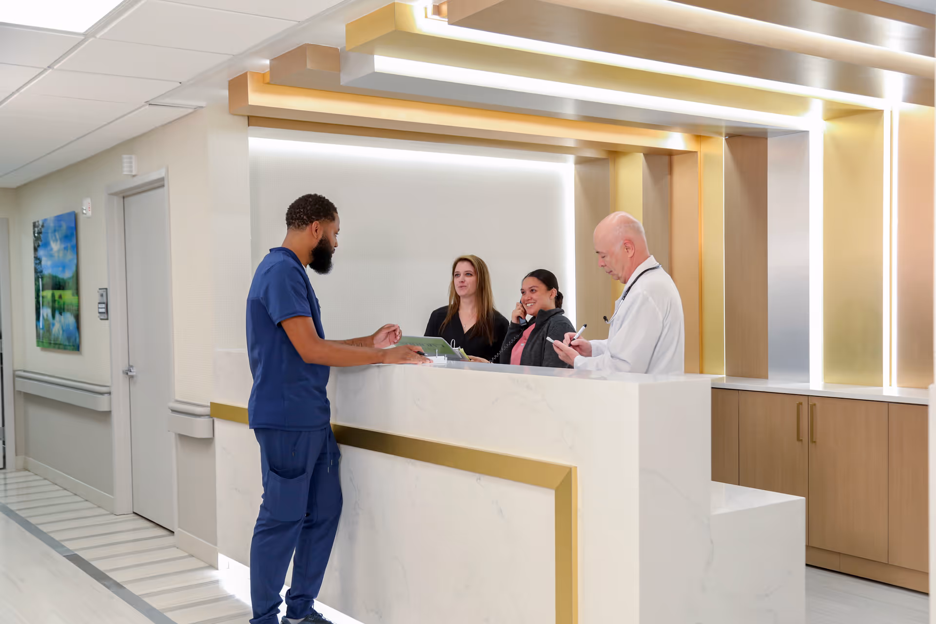 A modern reception area with a white and gold front desk where four people are interacting. One man in blue scrubs is standing on the visitor side of the desk, while two women and one man in a white coat stand behind the desk. The background features warm lighting and wooden cabinetry.