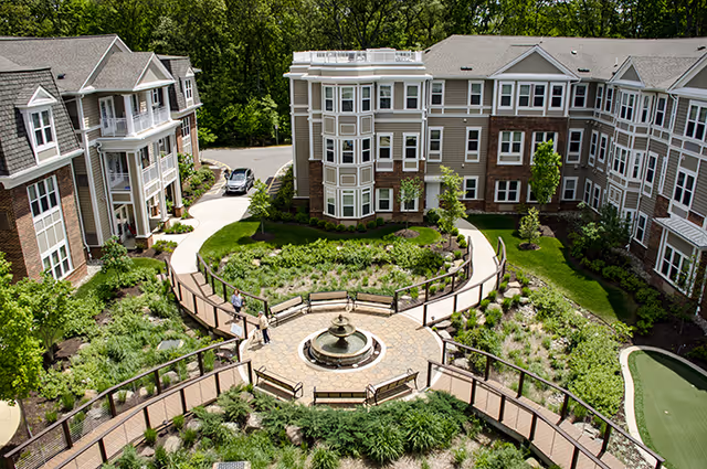 Aerial view of a landscaped courtyard with a central fountain, circular walkways and benches surrounded by multi‑story senior living buildings and trees.