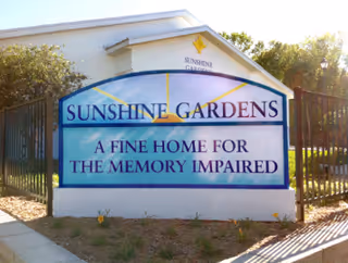 Outdoor sign for Sunshine Gardens, a facility described as 'A fine home for the memory impaired,' with a building and trees in the background.