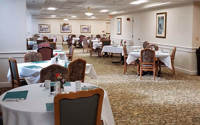 A spacious dining room in a senior living community with multiple round and square tables covered with white tablecloths. Each table is set with green placemats, white mugs, silverware, and small flower arrangements. The room has patterned carpet flooring, beige walls with framed artwork, and ceiling lights providing bright illumination.