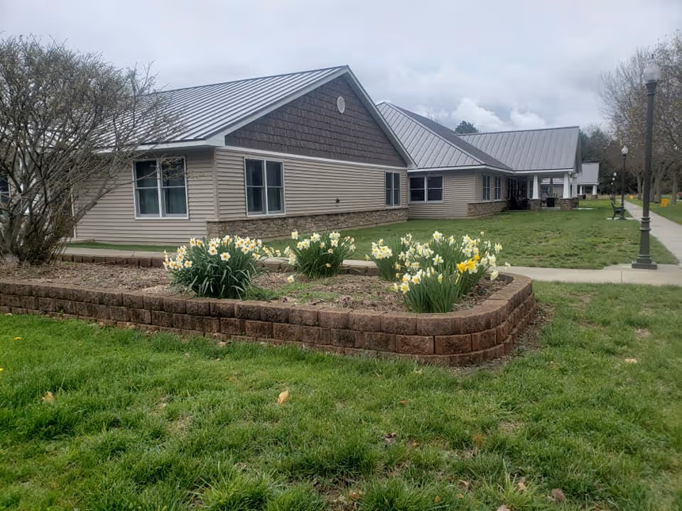 Exterior view of a single-story senior living facility building with beige siding and a metal roof. In front of the building is a raised flower bed with blooming white and yellow daffodils. A sidewalk runs alongside the building with street lamps and benches visible. The sky is overcast.