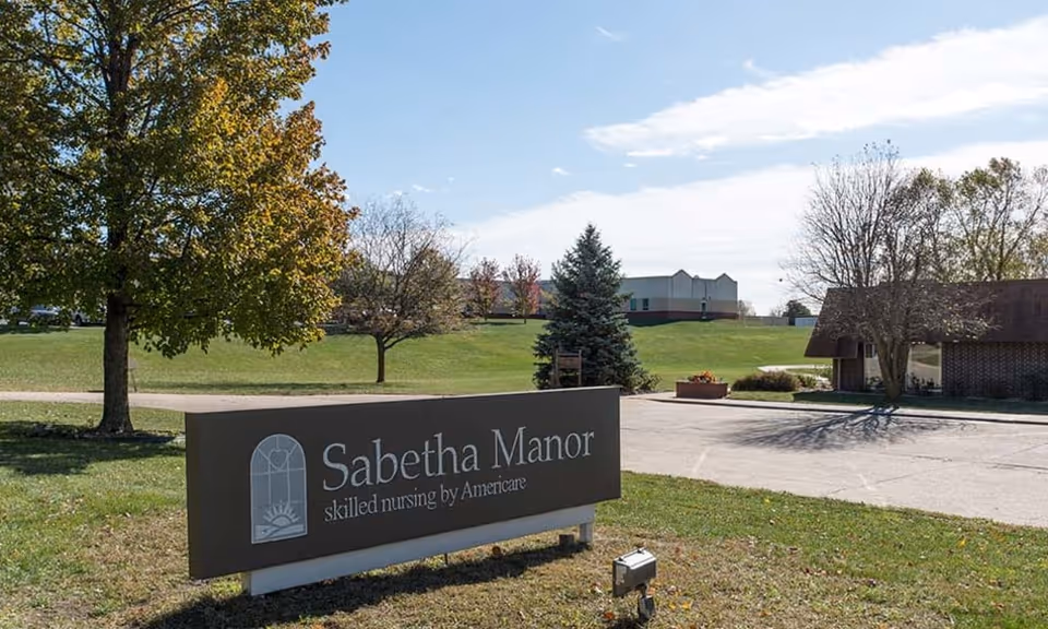Entrance sign reading 'Sabetha Manor skilled nursing by Americare' on a grassy lawn with trees and buildings in the background.