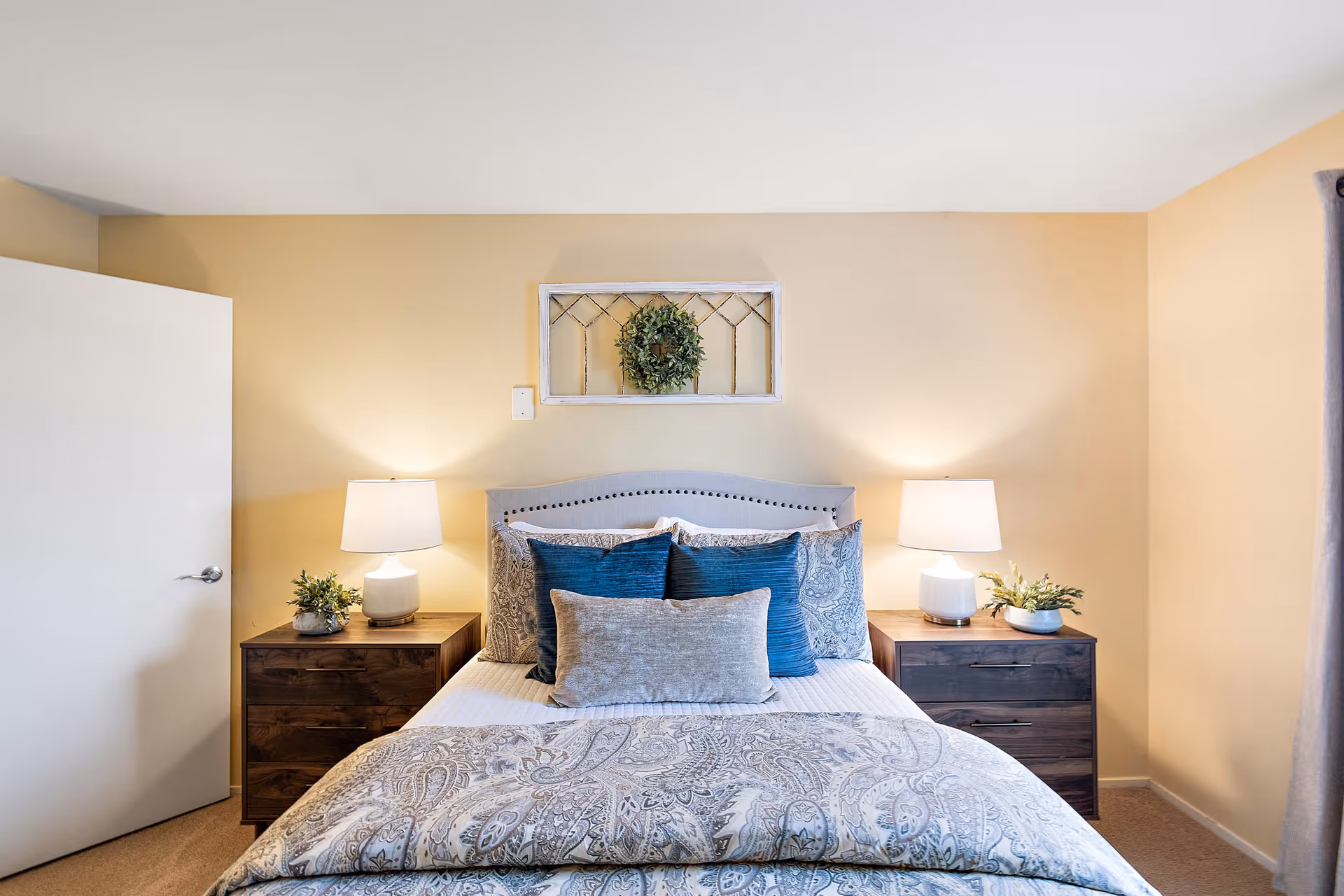 A neatly made bed with patterned bedding and multiple pillows in a bedroom. There are two wooden nightstands on either side of the bed, each with a white lamp and a small plant. A decorative wreath hangs on the wall above the bed. The walls are painted a soft beige color and a door is partially open on the left side of the room.
