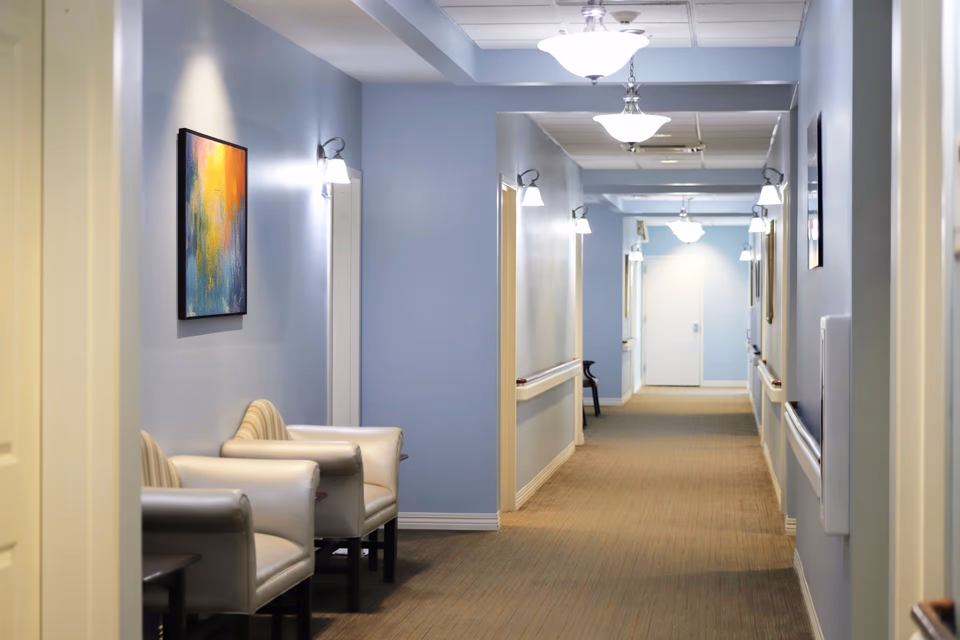 Well-lit hallway with pale blue walls, armchairs, wall sconces and artwork leading to a distant door.