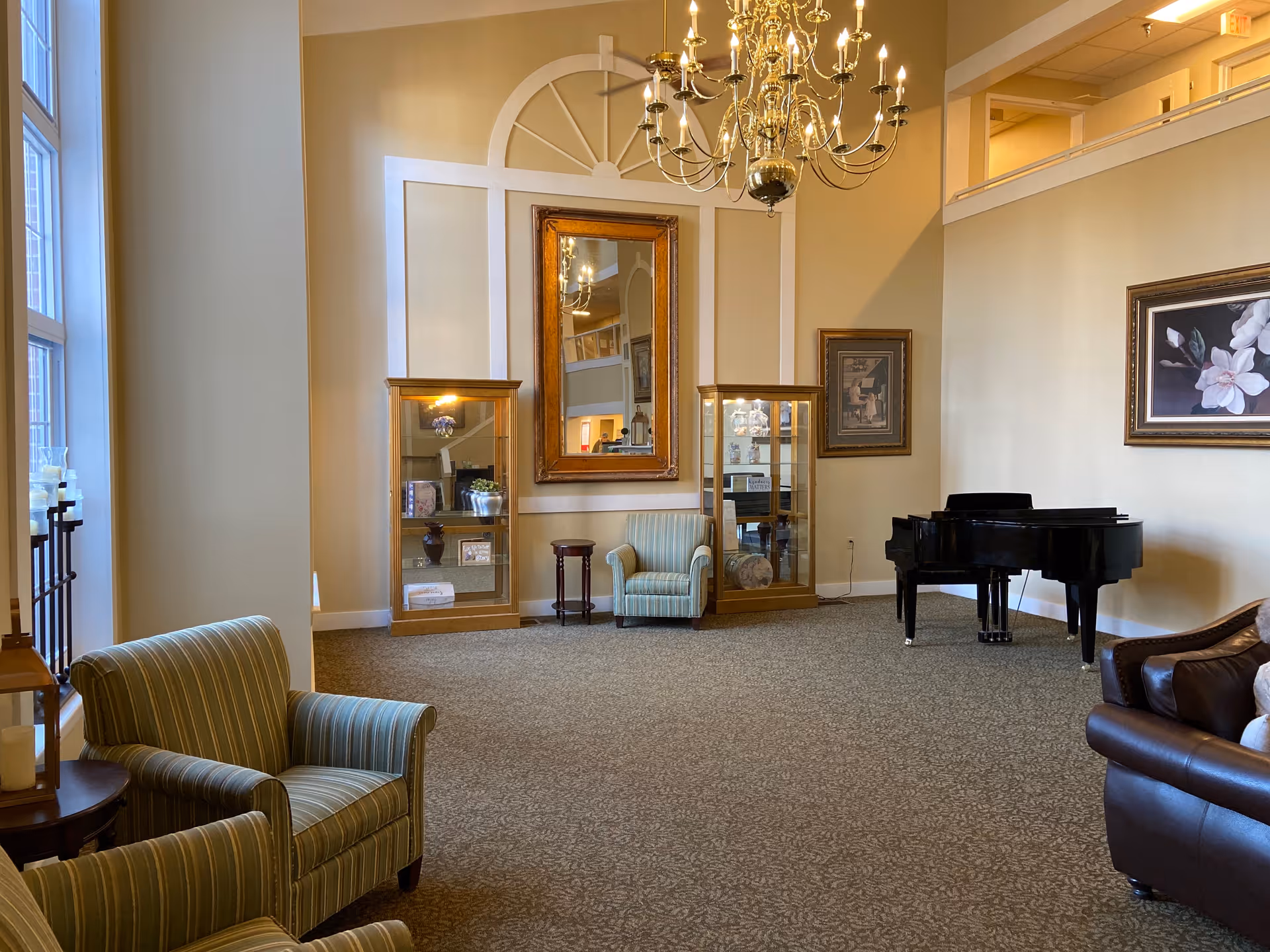 Spacious senior living lounge with upholstered armchairs, a grand piano, display cabinets, a large mirror and chandelier.