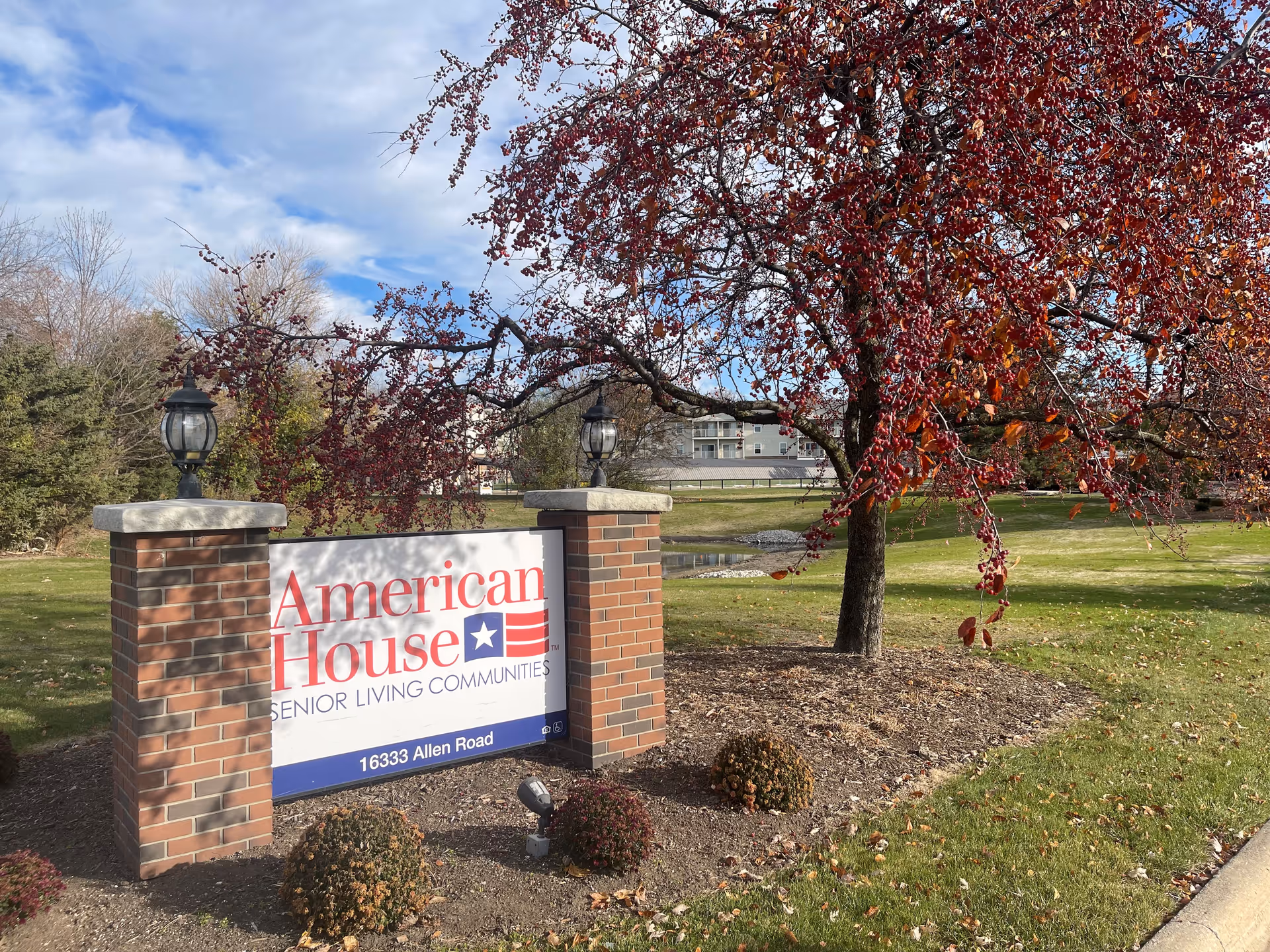 Brick entrance sign reading "American House Senior Living Communities" on a landscaped lawn with a tree and the facility building in the background.