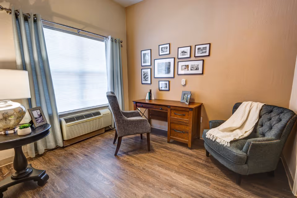 A cozy interior room with wooden flooring featuring a wooden desk with a gray upholstered chair in front of it. On the wall above the desk are multiple framed black and white photographs arranged in a gallery style. To the right of the desk is a dark gray tufted armchair with a cream-colored throw blanket draped over one arm. To the left, there is a round side table with a lamp, a small plant, and a framed photo. A large window with light blue curtains and blinds is behind the desk and chair, and below the window is an air conditioning unit.