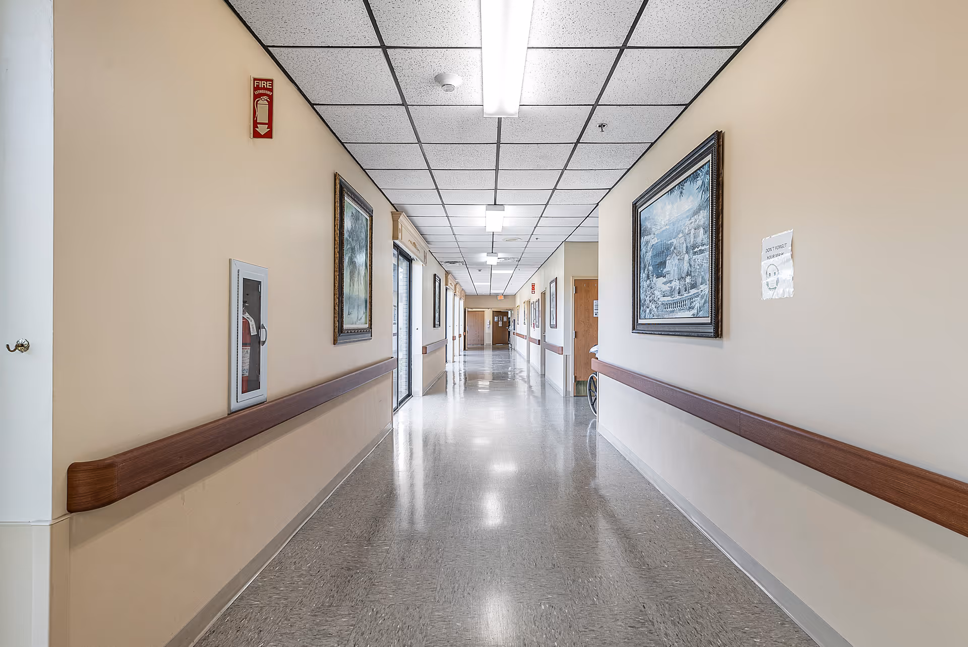 A long, clean hallway in a senior living facility with beige walls, wooden handrails on both sides, framed paintings hanging on the walls, and a fire extinguisher cabinet. The ceiling has fluorescent lights and a fire alarm. The floor is polished and reflective.