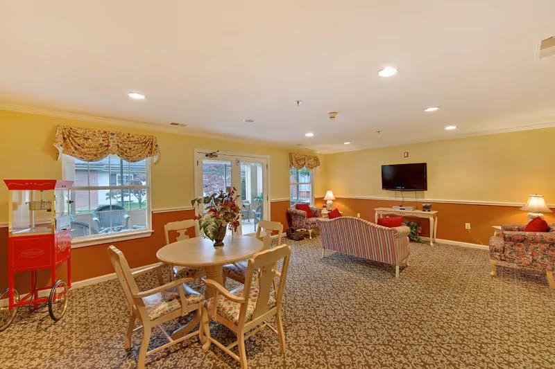 A cozy senior living common area with patterned carpet, a round wooden table with four chairs, floral curtains on windows, a red popcorn machine, several upholstered armchairs and a loveseat, a flat-screen TV mounted on the wall, and warm yellow and brown walls with lamps providing soft lighting.