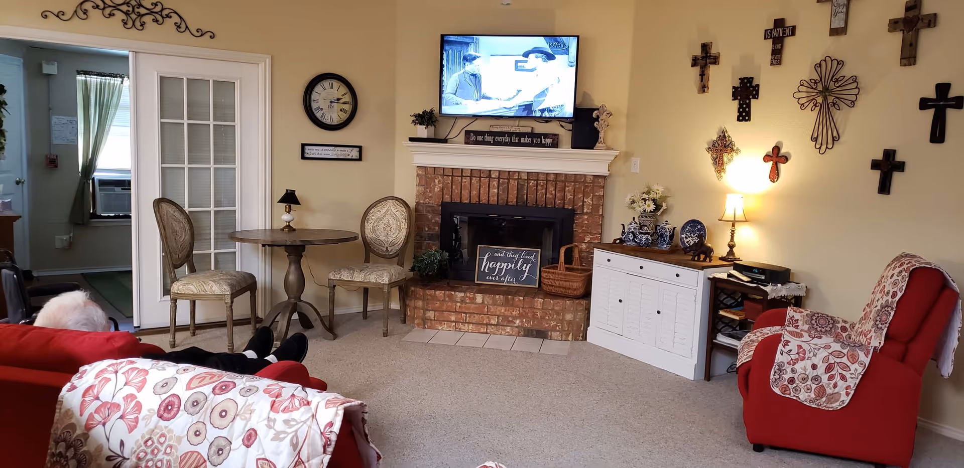 A cozy senior living room with a brick fireplace, a wall-mounted TV showing a black and white program, and various decorative crosses on the wall. The room has two red armchairs with floral patterned throws, a small round wooden table with two upholstered chairs, a white cabinet with blue and white ceramics, and a small lamp. An elderly person is seated on one of the armchairs.