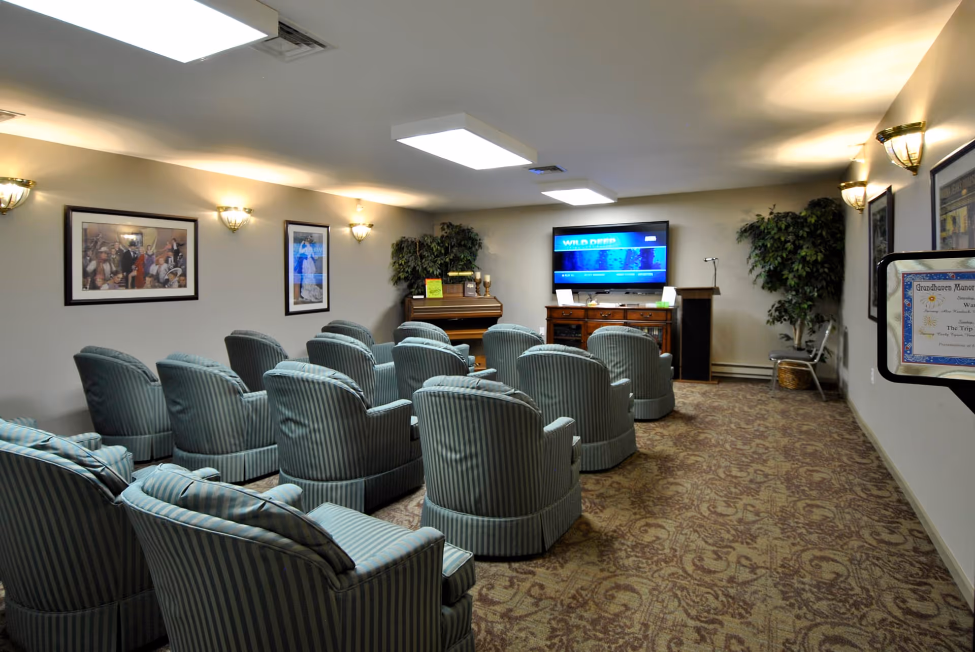A cozy common room with multiple striped armchairs arranged in rows facing a flat-screen TV mounted on the wall. The room has soft lighting from wall sconces, framed artwork on the walls, a piano in the corner, and a couple of potted plants. The carpet has a patterned design, and there is a small podium near the TV.